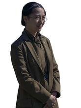 A woman wearing a dark brown tailored blazer over a matching collared top, with glasses and her hair pulled back, shown against a plain white background.