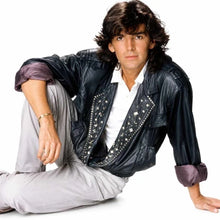 A man wearing a black leather jacket with silver studs and a white shirt, sitting on a white background.