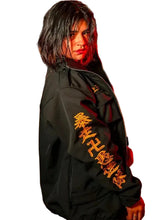 A person standing outdoors at night wearing a black jacket with large gold Japanese characters printed down one sleeve. The background shows a blurred city street scene with warm orange lighting and streetlights.