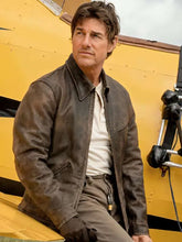 A man wearing a brown leather jacket, white shirt, and khaki pants sits against a yellow aircraft body.