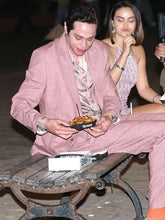 A man in a pink suit and patterned shirt sits on a bench holding a bowl of food, with a woman in a light pink dress seated behind him in a dimly lit setting.