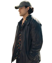 A man wearing a black leather jacket over a dark floral shirt and a dark baseball cap, standing outdoors on a sunny street with trees and buildings in the background.