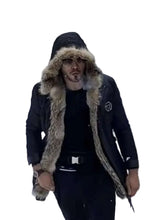 A man wearing a black winter parka with a thick beige fur-trimmed hood, standing on a snowy road with snow-covered pine trees in the background.