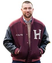 A man wearing a maroon and black varsity jacket with 'New York City' and 'HILTI' embroidery, standing outdoors with a blurred green background.