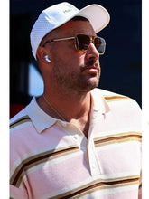 A man wearing a light pink and white striped polo shirt, a white baseball cap, and aviator sunglasses, with a white wireless earbud visible in his ear.