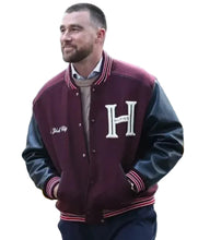 A man wearing a burgundy and black varsity jacket with a large white 'H' on the chest, standing in an outdoor city setting with yellow taxis and buildings in the background.