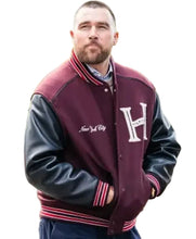 A man wearing a maroon and black varsity jacket with 'New York City' and a large 'H' on the chest, photographed outdoors in a park-like setting.