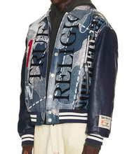 A model wearing a True Religion varsity-style jacket with a blue and gray denim collage print on the body and navy leather sleeves, featuring 'TRULOGIC' text and striped ribbed cuffs.