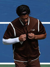 A man on a blue tennis court wearing a brown Nike jacket with white trim over a white long-sleeve shirt and brown pants, adjusting the front of the jacket.