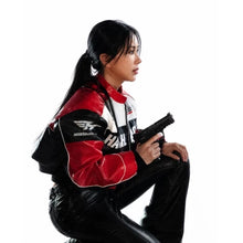 A woman in a red, black, and white racing jacket and black pants crouches against a white background, holding a black toy gun. The jacket features the visible brand name 'AKUMU' on the sleeve.