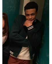 A young man wearing a dark green varsity jacket with white stripes on the cuffs and hem, layered over a cream sweatshirt and brown pants, standing against a teal wall.