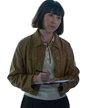 A woman with a short dark bob haircut is shown from the waist up, wearing a brown utility jacket over a cream shirt. She is holding a clipboard and pen against a solid white background.