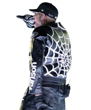 A performer on stage seen from the back wearing a black cap, a black jacket with a large white spiderweb graphic across the back, and dark pants, with a stadium crowd in the background.