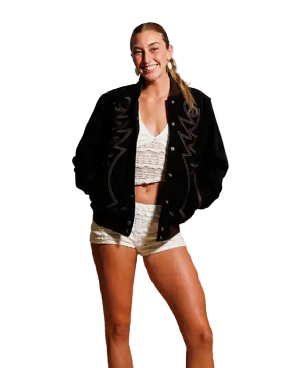 A woman posing on wooden steps wearing a black varsity-style jacket with white embroidered lettering, a white lace crop top, and white shorts against a concrete wall.