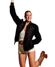 A woman posing in a black varsity-style bomber jacket with pink embroidered lettering, paired with a white lace crop top, white shorts, and brown cowboy boots against a concrete wall and wooden bench.