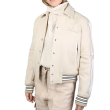 A model wearing a cream-colored varsity-style jacket with white sleeve stripes, layered over a cream knit turtleneck sweater and matching cream pants, against a solid gray background.