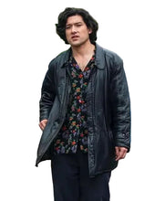 A man wearing a black leather jacket over a dark floral button-up shirt and black trousers, standing on a city street with trees, pedestrians, and a white vehicle in the background.