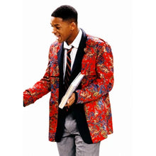 A man wearing a vibrant red patterned blazer with black lapels, a white shirt, a striped tie, and gray trousers, holding a book against a plain white background.