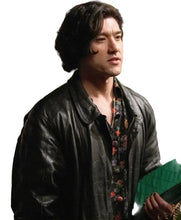 A man wearing a black leather jacket over a floral button-down shirt, holding a green textured clutch bag in an outdoor setting.