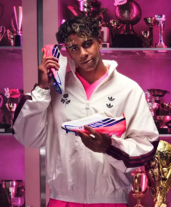 A man wearing a white Adidas track jacket and pink athletic shorts holds a pink and blue soccer cleat in a trophy-filled room with pink lighting. He is holding a pink phone to his ear.