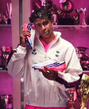 A man wearing a white Adidas track jacket and pink athletic shorts holds a pink and blue soccer cleat in a trophy-filled room with pink lighting. He is holding a pink phone to his ear.