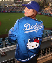 A woman at a baseball stadium wearing a shiny blue Los Angeles Dodgers jacket with a Hello Kitty graphic on the back, paired with a matching blue cap.