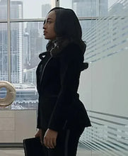 A woman in a black tailored blazer and matching trousers stands in a modern office interior with large windows showing a city skyline. She is shown in profile holding a dark folder or briefcase.