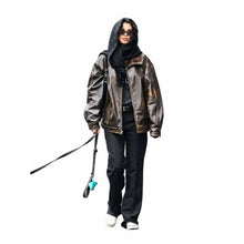 A model wearing a metallic bronze-brown leather bomber jacket, black hoodie, black pants, and white sneakers, holding a black leash with a blue tip against a solid white background.