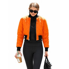A woman modeling a bright orange quilted bomber jacket over a black top and black pants, holding an iced coffee and a black handbag against a plain white background.