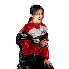 A woman wearing a red, white, and black motorcycle racing jacket with 'HARSH' and 'MotoGP' graphics, holding a black toy gun against a solid white background.