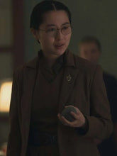 A woman wearing round wire-frame glasses and a brown tailored jacket over a dark top, holding a small round object in a dim indoor setting.