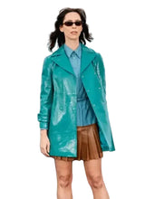 A woman wearing a shiny teal-green raincoat over a blue shirt and brown pleated skirt, standing outdoors with a cityscape background and a black event backdrop featuring Hulu, Disney+, and FX logos.