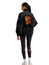 A model shown from the back wearing a black windbreaker jacket with a large orange graphic on the back, paired with black leggings and white sneakers, against a light gray studio background.