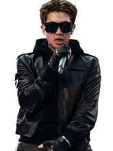 A man wearing a black leather jacket, black sunglasses, and black gloves, holding a microphone against a solid white background.