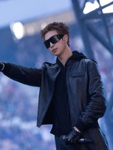A man on a stage wearing a black leather jacket, black sunglasses, and a black hoodie, holding a microphone with one hand while extending the other out.