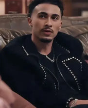 A man wearing a black zip-up jacket with silver studs and chain details, sitting on a brown leather couch.