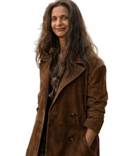 A woman with long wavy brown hair stands outdoors wearing a brown suede-like double-breasted coat over a dark top. The background shows a rustic white fence, parked vehicles, and bare trees in a field.