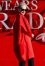 An older woman with gray hair and sunglasses stands in front of a bright red backdrop with large white lettering, wearing a voluminous bright red coat or cape and black pants.