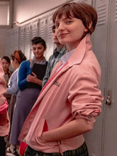 A person in a school hallway wearing a light pink jacket with 'Bella' embroidered on the chest, standing in front of gray lockers with other people blurred in the background.